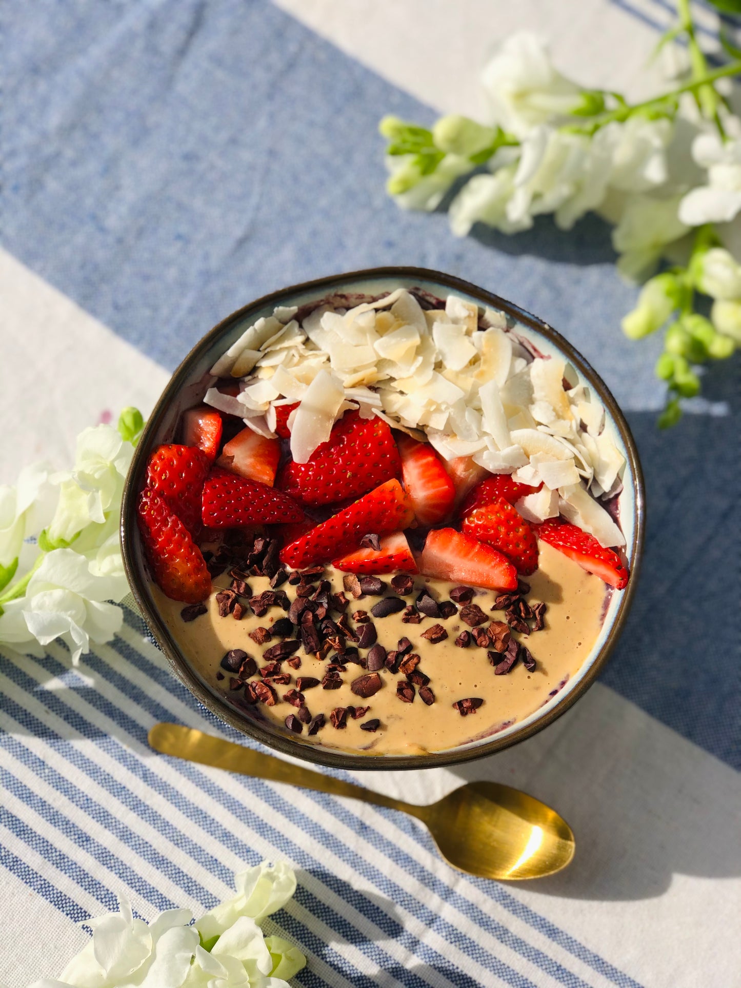 Healthy Peanut Butter breakfast bowl with granola, peanut butter, cacao nibs, strawberries and desiccated coconut  on a blue and white checked table, with a gold spoon and some flowers surrounding the bowl. 